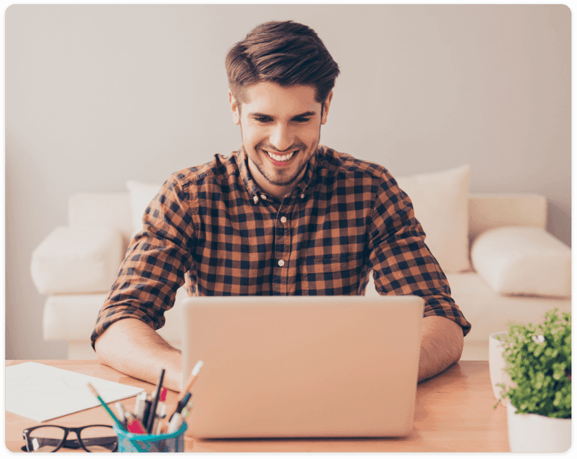 Homem jovem com uma camisa xadrez sorrindo enquanto trabalha em seu laptop em um ambiente de escritório doméstico com um sofá ao fundo e utensílios de escritório à frente